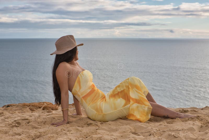 joven-y-bonita-mujer-cabello-negro-con-un-vestido-amarillo-sombrero-mirando-al-mar-sentado-en-...jpg